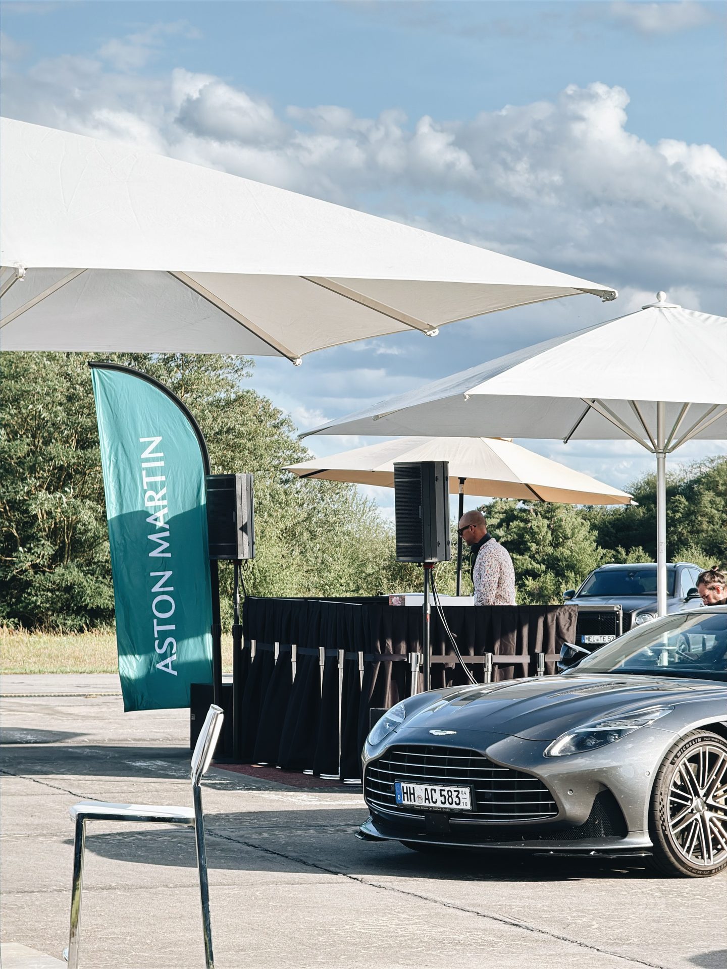 Aston-Martin-Flagge, DJ-Bereich und grauer Sportwagen unter Sonnenschirmen auf dem Flugplatz