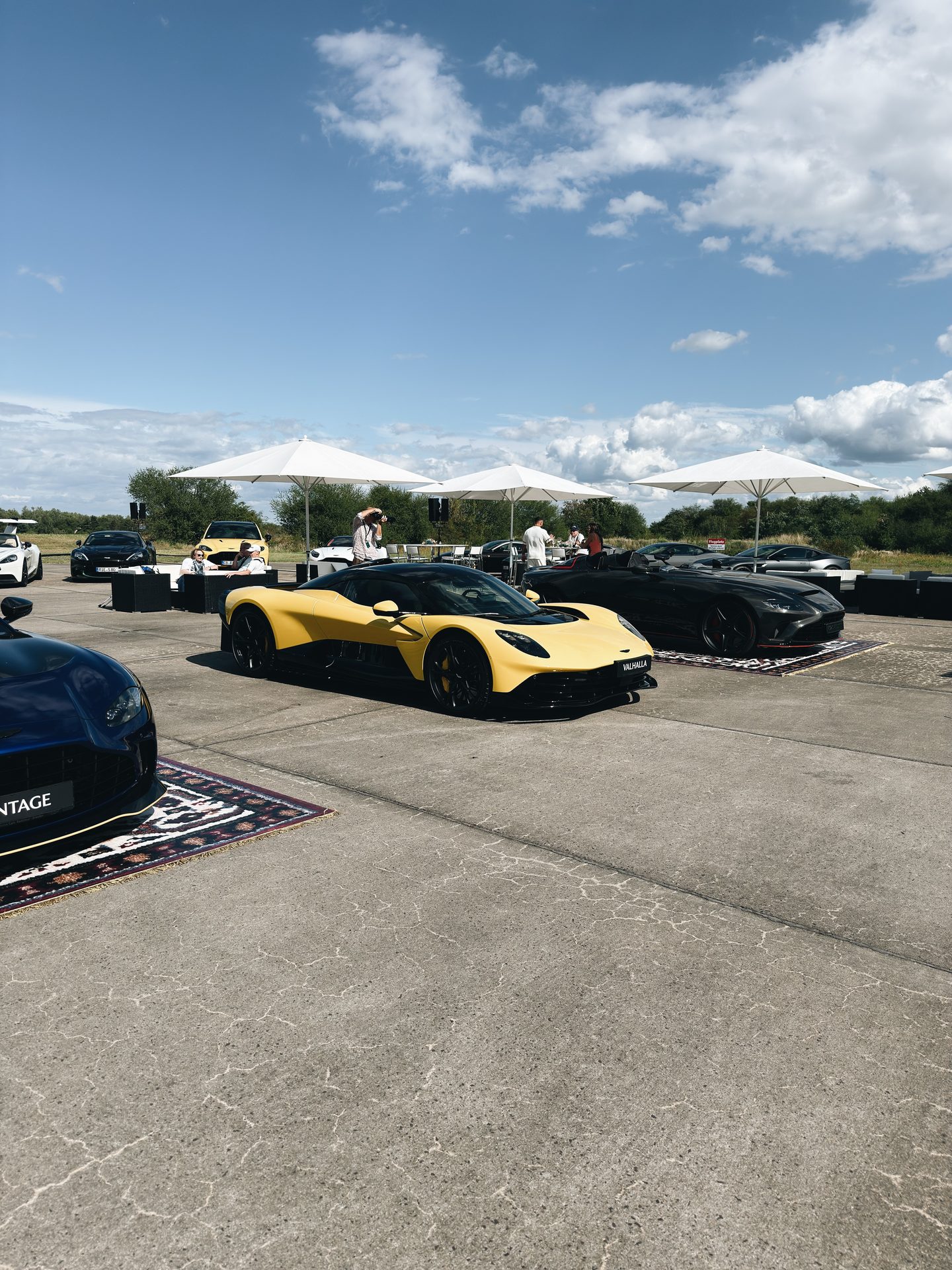 Gelber Aston Martin Valhalla und schwarzer Speedster auf Teppichen präsentiert unter weißen Sonnenschirmen